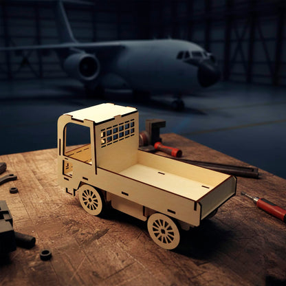 Laser cut wooden  plywood utility truck model on a workbench in an airplane hangar. Simple plywood 3D puzzle with an open bed and window mesh detail. Features spoke-style wheels and dark burnt edges, shown with an aircraft in the background.