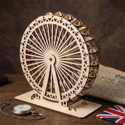 3D laser cut plywood model of the London Eye Ferris wheel shown at an angle. The natural wood grain and dark burnt edges are visible. It stands on a rectangular wooden base next to a silver pocket watch and a Union Jack flag for scale.