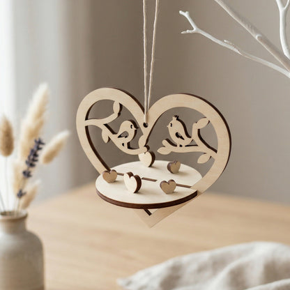Close-up of a 3D laser-cut plywood heart ornament featuring two birds perched on branches. The design includes miniature standing hearts on a circular base and a dark-edged natural wood finish. Hanging by a simple twine string in a bright, minimalist setting.