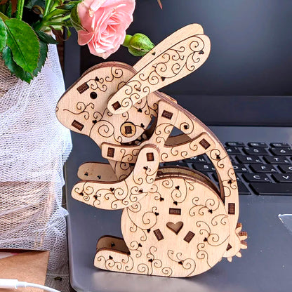 A highly decorative laser cut wooden plywood rabbit figure with heart engravings sits on a silver laptop, next to a vase of pink miniature roses and white wireless earphones