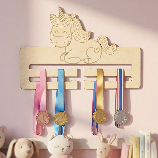 Laser cut plywood medal holder featuring a cute sitting unicorn with a heart on its hip and a striped horn. The light wood display has four horizontal slots for hanging ribbons, mounted on a pink wall above a shelf with plush toys.