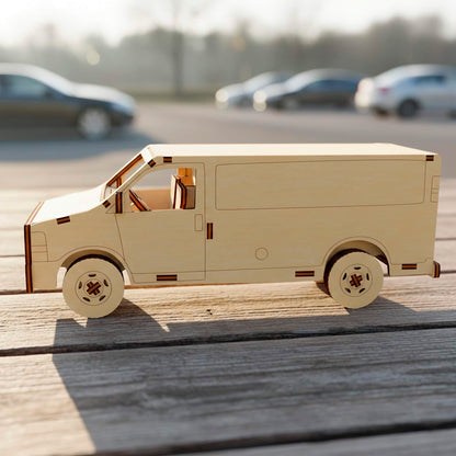 Side view of a laser cut wooden plywood delivery van model on a rustic wooden surface. The 3D plywood puzzle features clean lines, detailed wheels, and visible assembly joints, with a blurred outdoor parking lot background in daylight.