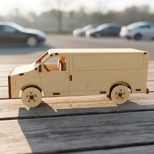 Side view of a laser cut wooden plywood delivery van model on a rustic wooden surface. The 3D plywood puzzle features clean lines, detailed wheels, and visible assembly joints, with a blurred outdoor parking lot background in daylight.