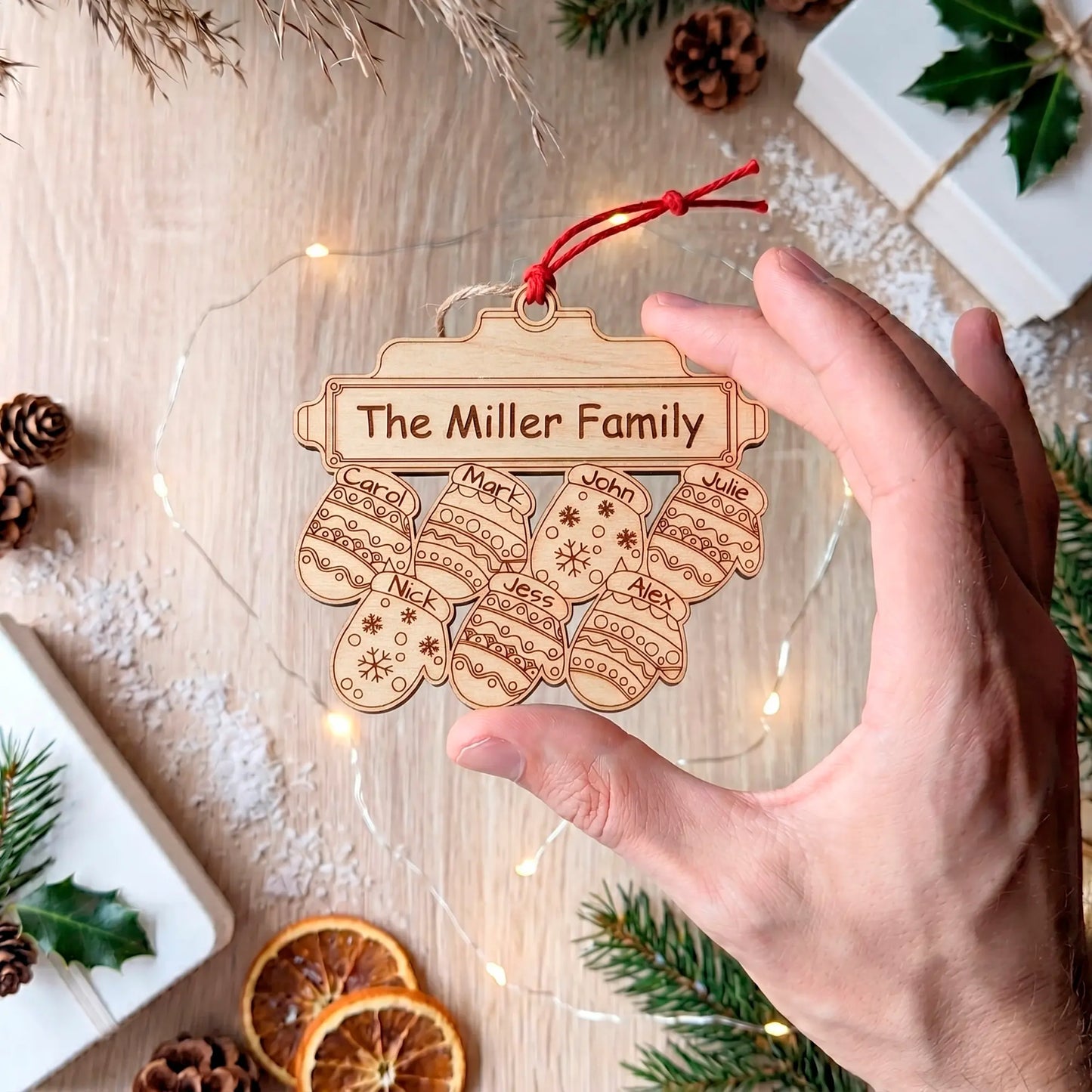 A hand holds an unpainted laser cut plywood ornament with 7 engraved, named mittens below "The Miller Family" plaque.