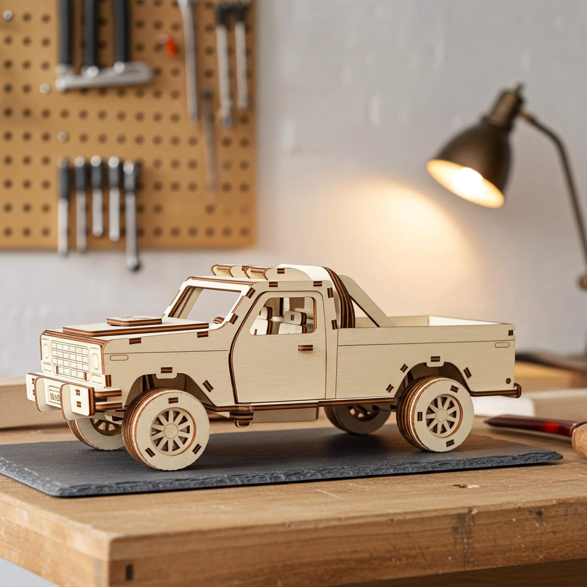 Laser cut wooden plywood pickup truck 3D model on a slate mat in a workshop. Detailed plywood assembly featuring a truck bed with a roll bar, off-road tires, and interior seats. Background shows a tool pegboard and a warm glowing desk lamp.