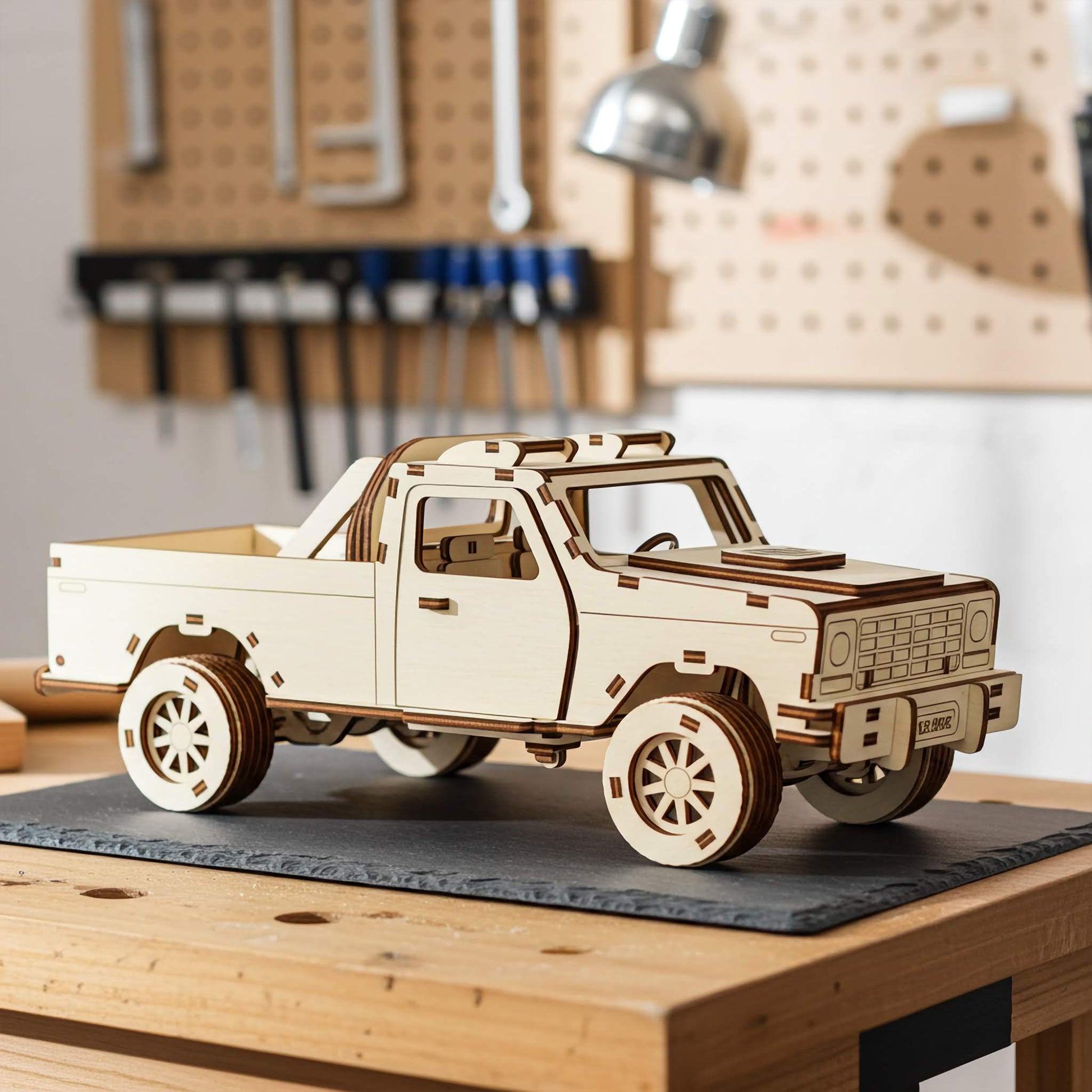 Laser cut wooden plywood pickup truck 3D model on a slate mat in a workshop. Detailed plywood assembly featuring a truck bed with a roll bar, off-road tires, and interior seats. Background shows a tool pegboard and a warm glowing desk lamp.