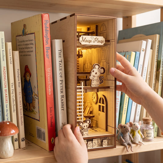 Hands touching a lit laser-cut birch plywood mouse house book nook on a shelf, highlighting wood grain and burnt edges.
