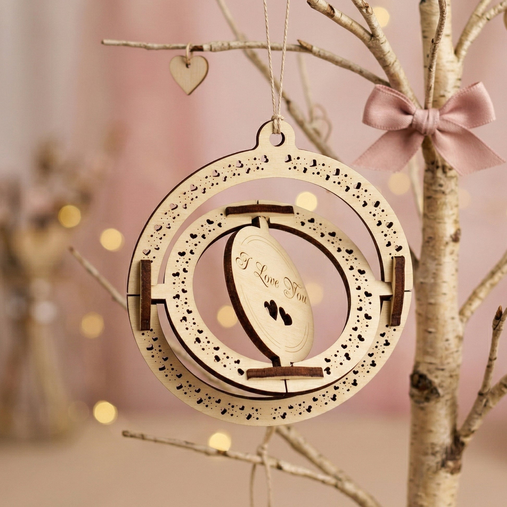 Side-angled view demonstrating the spinning mechanism of a 3D laser-cut plywood Valentine's card. The inner circle is tilted to show the kinetic design. Engraved with "I Love You" and heart patterns, the ornament is displayed on a decorative branch with a satin bow.