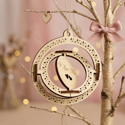 Side-angled view demonstrating the spinning mechanism of a 3D laser-cut plywood Valentine's card. The inner circle is tilted to show the kinetic design. Engraved with "I Love You" and heart patterns, the ornament is displayed on a decorative branch with a satin bow.