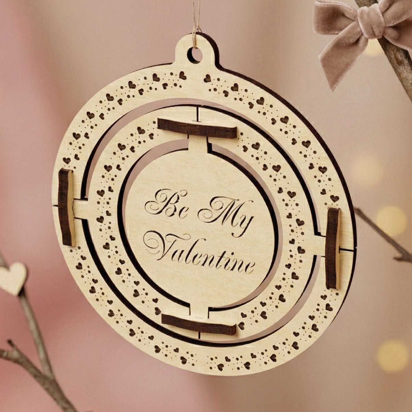 A 3D kinetic Valentine's Day ornament made of laser-cut plywood. The central circle is engraved with "Be My Valentine" in elegant script, surrounded by two nested moving rings with intricate heart patterns. Hanging from a branch with a soft velvet bow in a romantic, aesthetic setting.