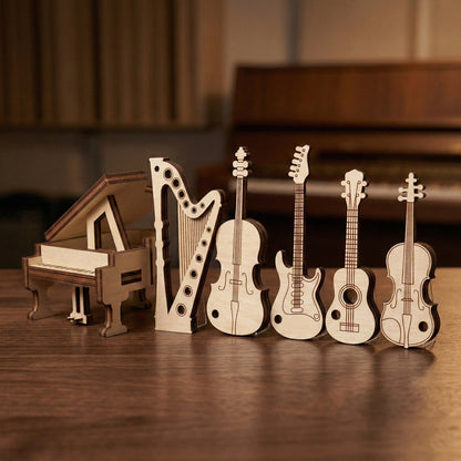 A row of laser cut plywood musical instrument miniatures standing upright, including a grand piano, harp, violin, electric guitar, acoustic guitar, and cello. A blurred piano and studio acoustic panels are visible in the background.