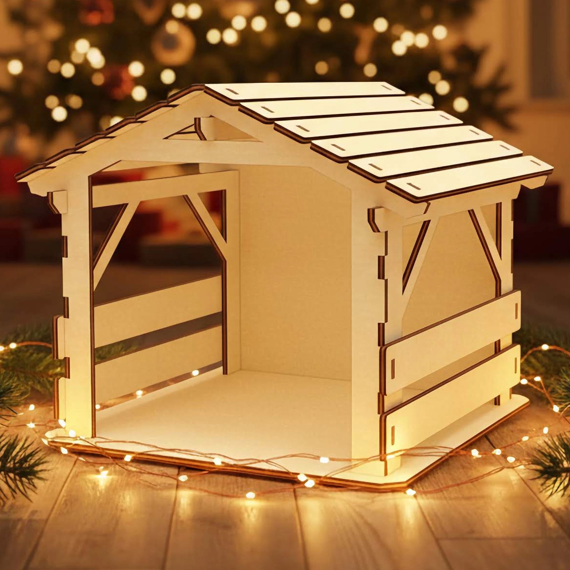 
Wooden laser cut Nativity barn #3 with classic slatted roof, open stable sides, and rustic beam details, softly illuminated against a blurred Christmas tree background.
