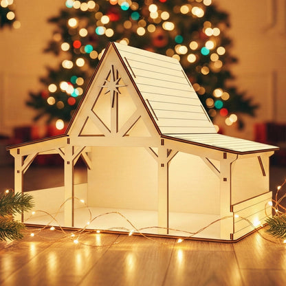 Wooden laser cut Nativity barn #4 with timber-framed open sides, extended roof overhang, and prominent star detail on the gable, warmly illuminated against a softly blurred Christmas tree background.