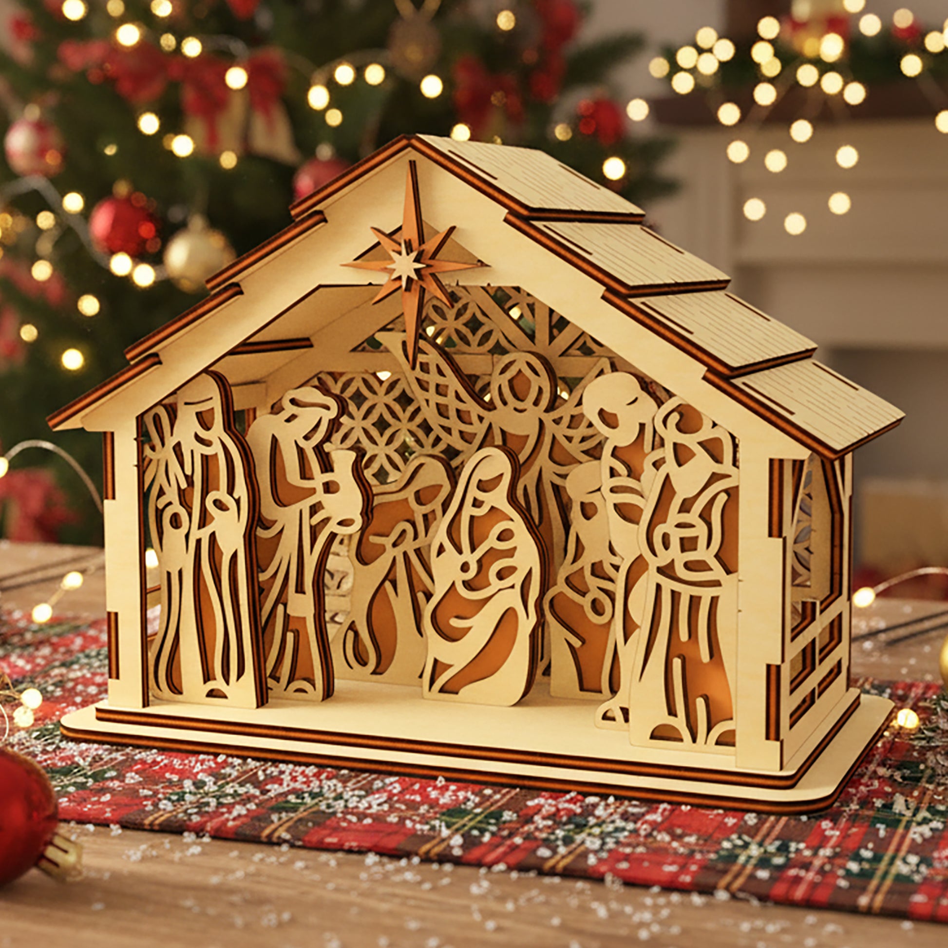Laser cut wooden 3D Nativity scene with layered Mary, Joseph, baby Jesus and Bethlehem star, illuminated from within against a warm Christmas tree bokeh background.