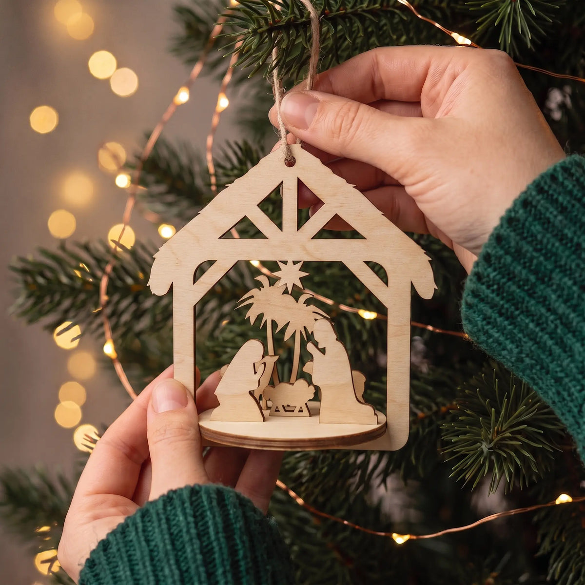 Hands hold laser cut plywood nativity creche bauble showing scale in front of warm Christmas tree lights.