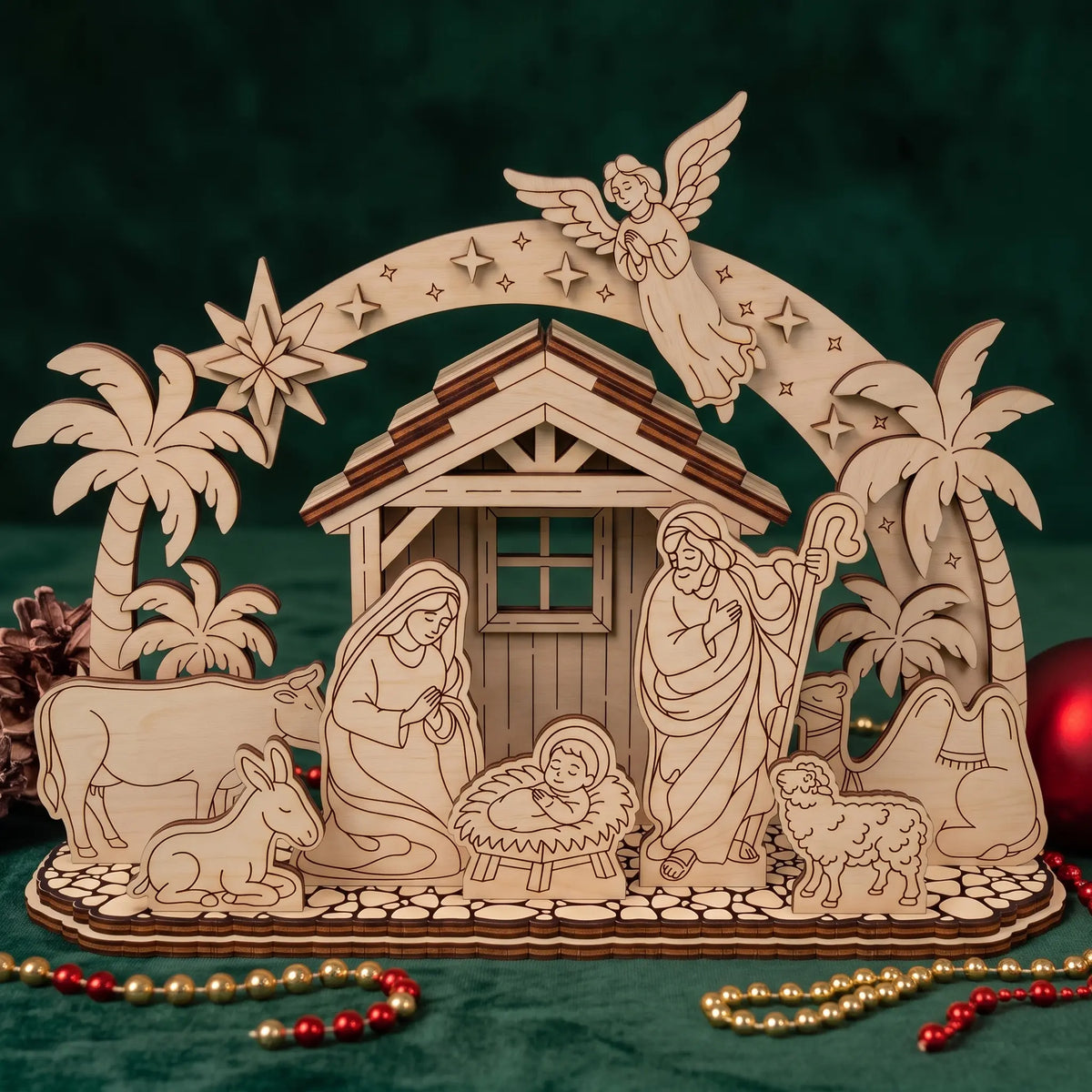 Laser cut plywood nativity set on green fabric with palm trees, angel arch, and holiday beads.