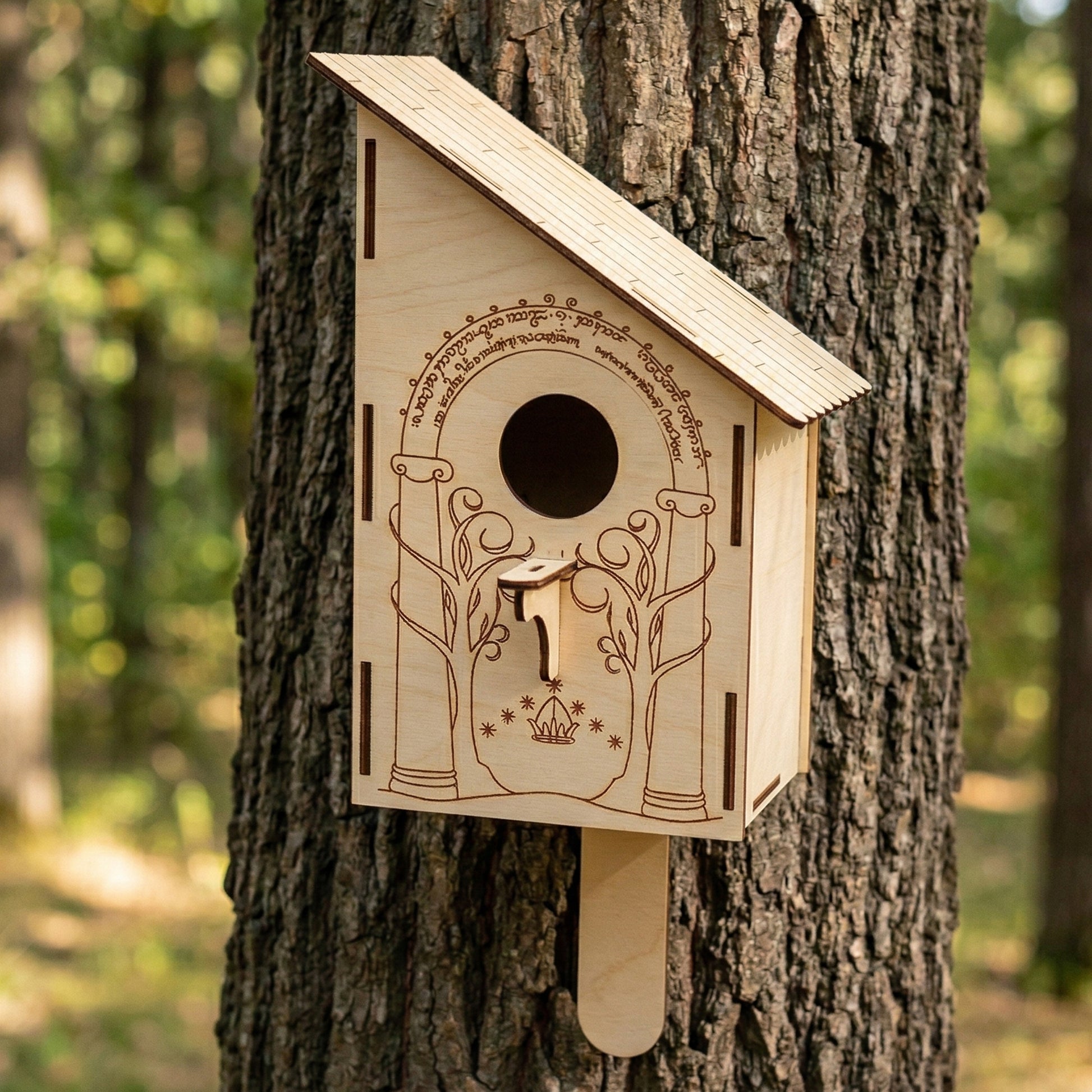A handcrafted laser-cut plywood nesting box featuring an intricate engraving of the Doors of Moria. The wooden birdhouse has a sloped shingle-style roof, a circular entrance hole, and a small perch. Mounted on a large tree trunk in a sunlit forest, showcasing the detailed Elvish-style script and fantasy-themed design.