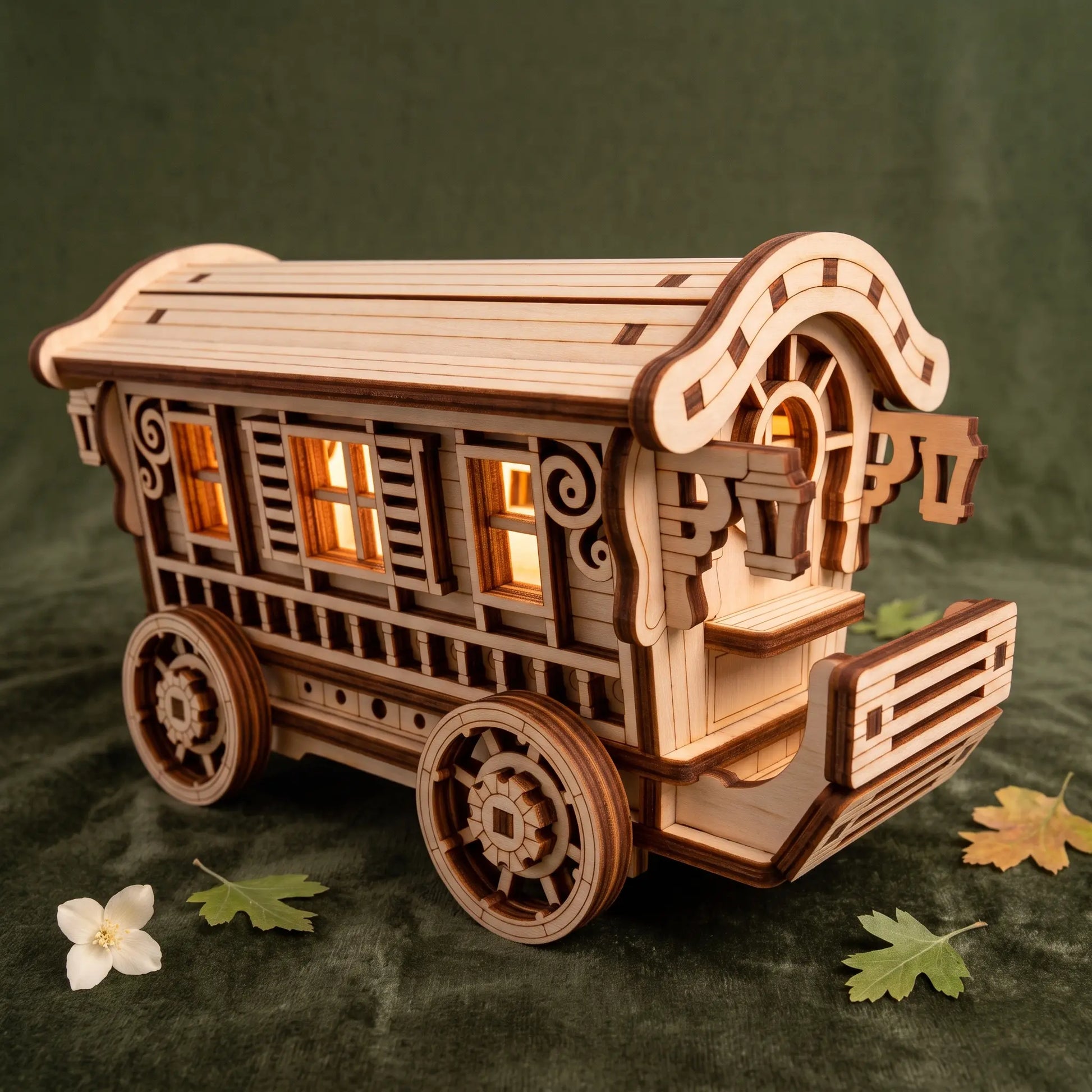 A wooden 3D laser-cut model of a traditional nomad wagon, illuminated from within by warm yellow lights, displayed on a dark green textured background with small decorative leaves and a flower.