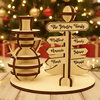 Wooden laser cut North Pole Family Sign featuring a smiling snowman and a personalized directional signpost with family names, displayed against a festive blurred Christmas tree background.
