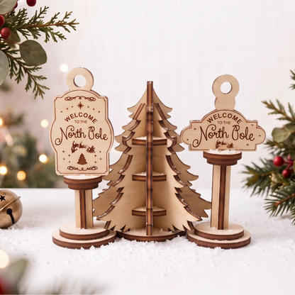 Laser cut plywood North Pole signs and slotted pine tree standing in artificial snow beside brass jingle bell and berries.