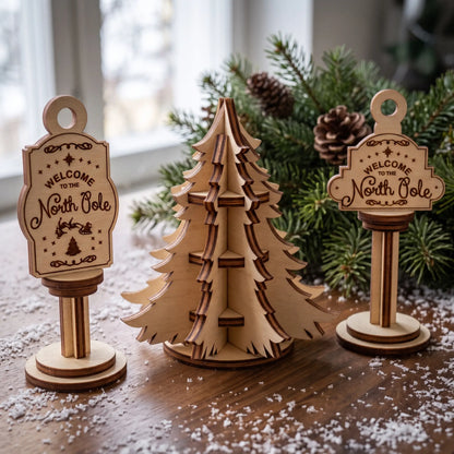 Set of laser cut plywood North Pole post ornaments and 3D mini tree on snowy table in front of lush green pine branches.