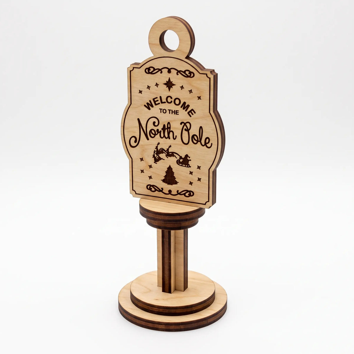 Front view of vertical laser cut plywood Welcome to the North Pole sign on round stand against plain white background.