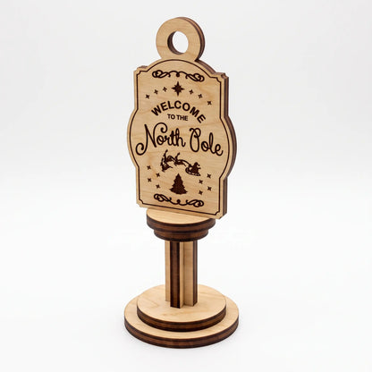 Front view of vertical laser cut plywood Welcome to the North Pole sign on round stand against plain white background.