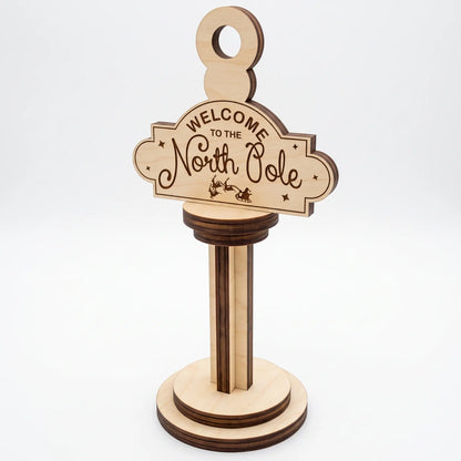 Horizontal laser cut plywood Welcome to the North Pole post ornament on stepped wooden base against white background.