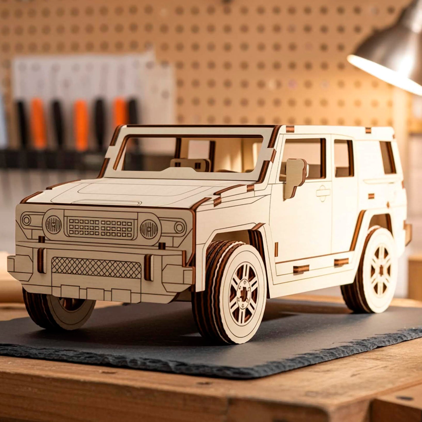 Laser cut wooden plywood 3D model sitting on a slate mat in a workshop setting. Detailed plywood puzzle assembly featuring a textured front grille, round headlights, and realistic tires. Background shows a pegboard with tools and a warm desk lamp.