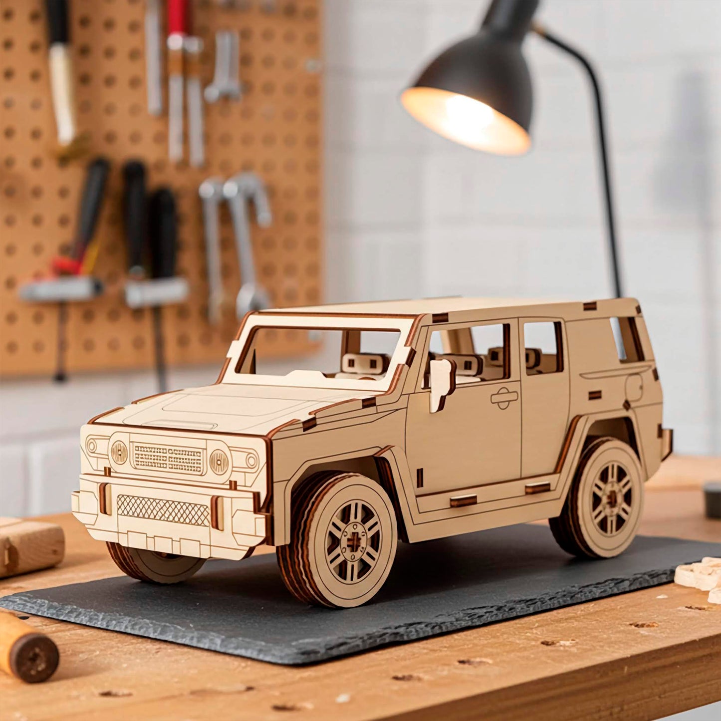 Laser cut wooden plywood 3D model sitting on a slate mat in a workshop setting. Detailed plywood puzzle assembly featuring a textured front grille, round headlights, and realistic tires. Background shows a pegboard with tools and a warm desk lamp.