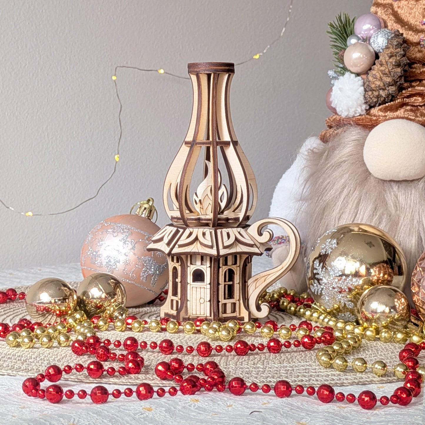 Laser-cut wooden fairy house shaped like an oil lamp, surrounded by Christmas ornaments and festive beads indoors