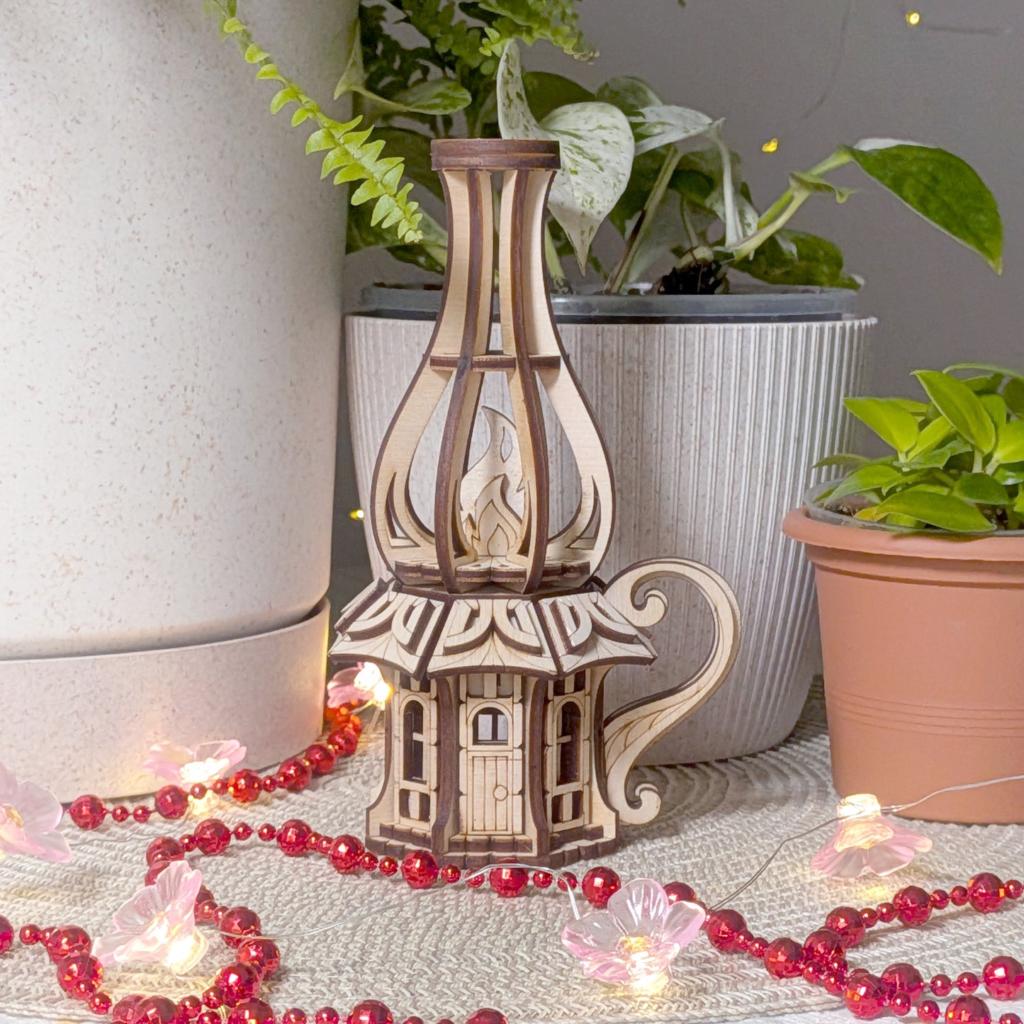 Decorative plywood oil-lamp fairy house placed near potted plants, showing detailed laser-cut flame and roof design