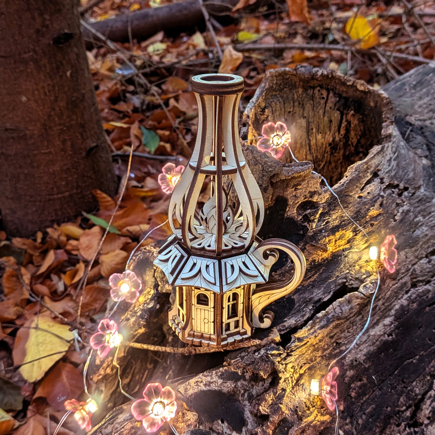 Laser-cut wooden oil-lamp fairy house displayed outdoors on bark with glowing flower lights and autumn leaves