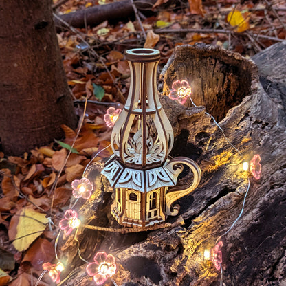 Laser-cut wooden oil-lamp fairy house displayed outdoors on bark with glowing flower lights and autumn leaves