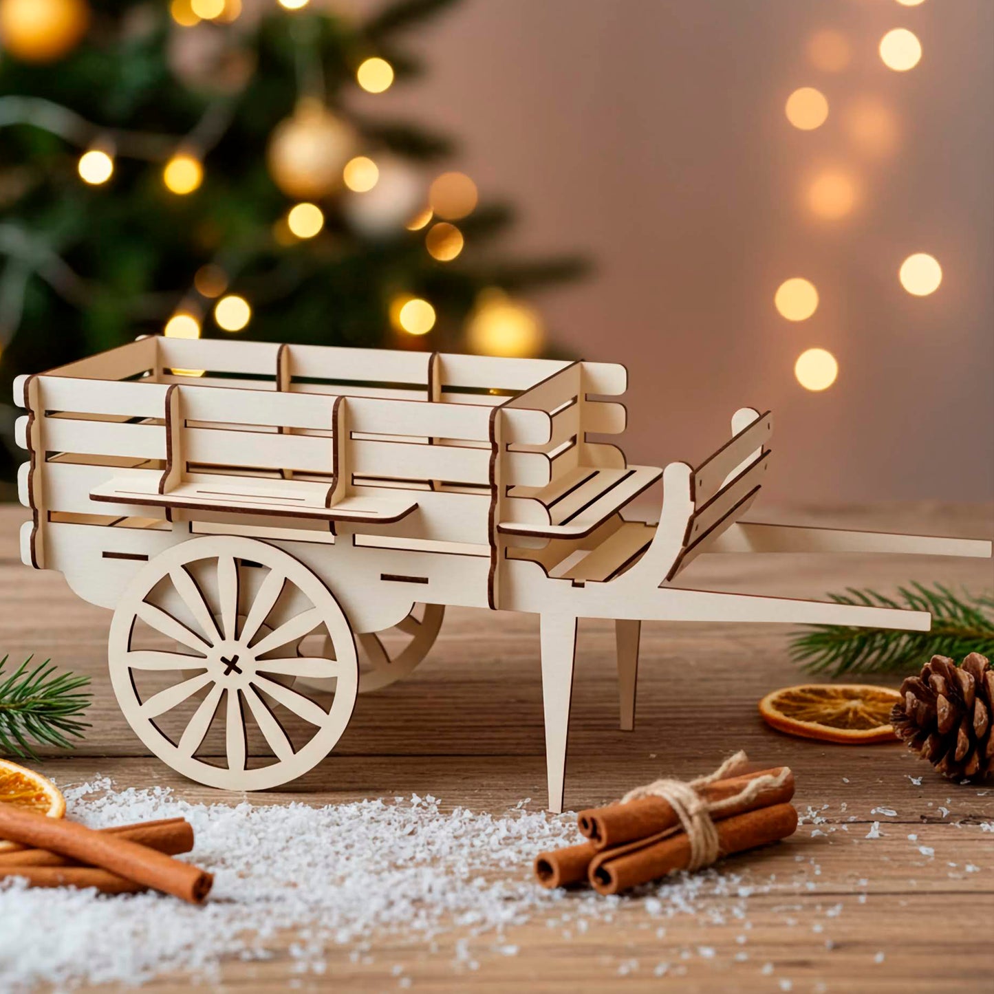A light brown laser cut wooden plywood miniature wagon sitting on a wooden table with faux snow, cinnamon sticks, and dried orange slices, with a glowing Christmas tree in the blurred background.