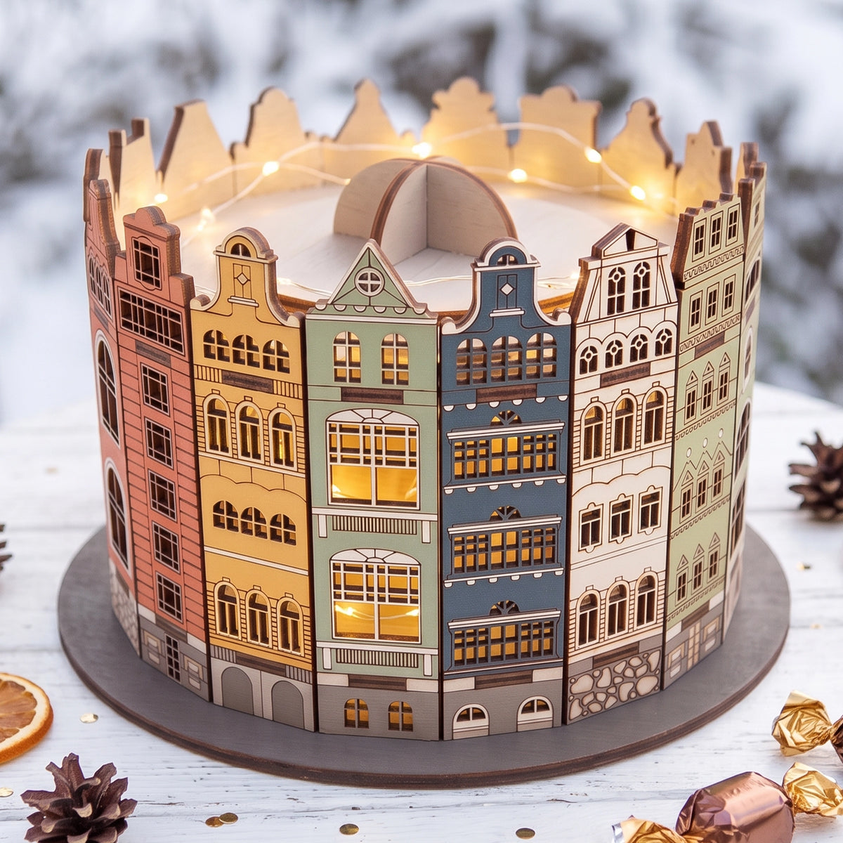 Illuminated laser cut plywood old town candy box glowing on snowy wooden table with pinecones, lights, and sweet treats.
