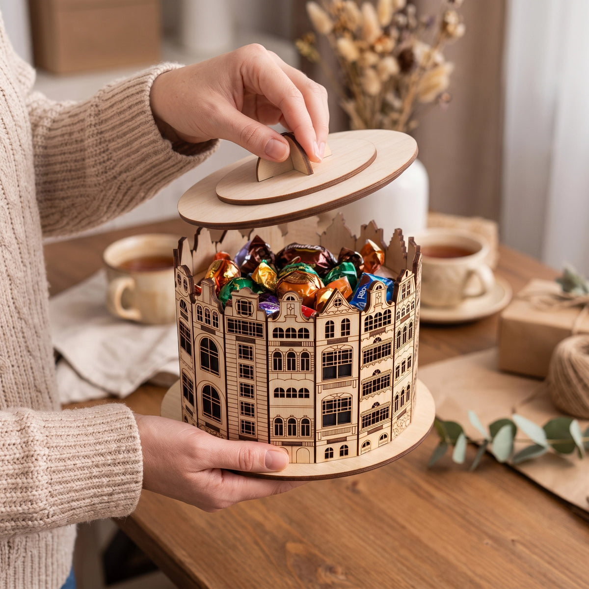 Woman opening a laser cut plywood old town candy box filled with wrapped chocolates on a cozy wooden tea table with cups.