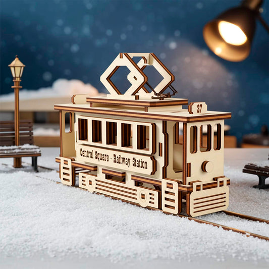 A light wooden plywood laser cut model of a vintage tram with the text "Central Square - Railway Station," sitting on snow-covered rails outdoors, with a street lamp and benches visible in the blue, snowy night background