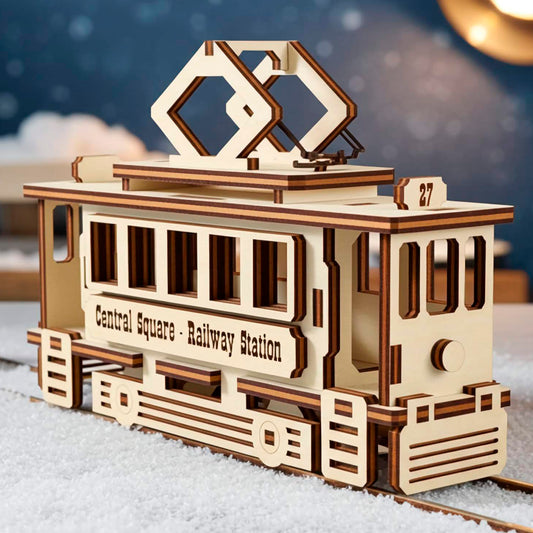 A light wooden plywood laser cut model of a vintage tram with the text "Central Square - Railway Station," sitting on snow-covered rails outdoors, with a street lamp and benches visible in the blue, snowy night background