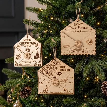 Three laser cut plywood holiday letter envelope ornaments hanging on Christmas tree with festive lights.