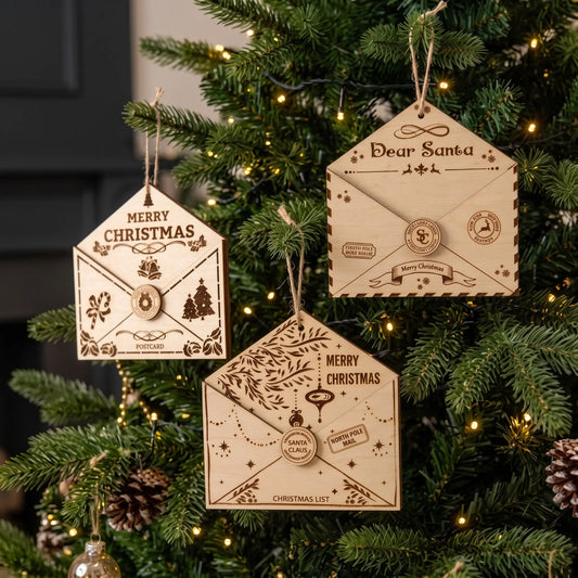Three laser cut plywood holiday letter envelope ornaments hanging on Christmas tree with festive lights.