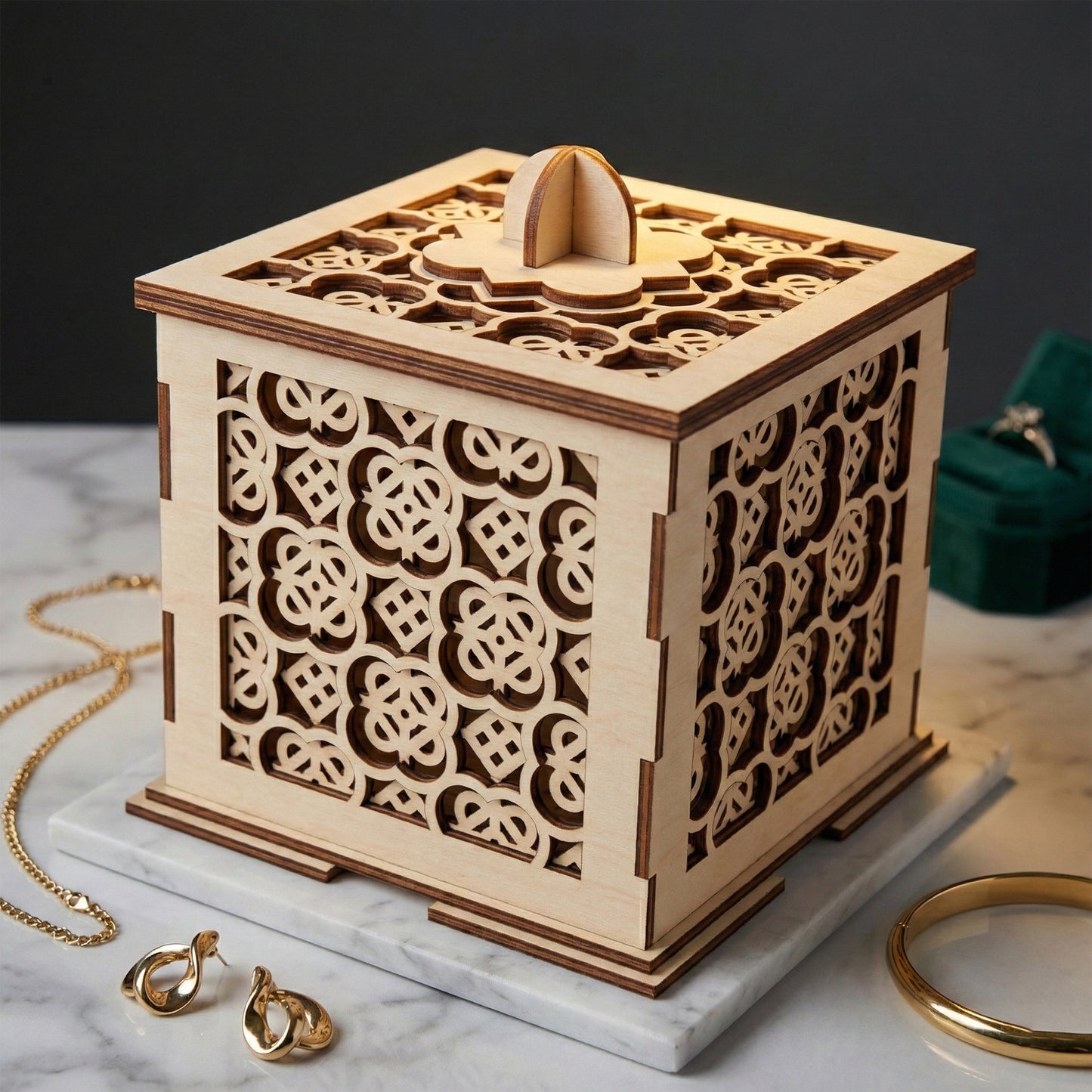 Square laser cut plywood jewelry box with intricate geometric filigree patterns on all sides and the lid. A small rounded wooden handle sits on top. The box is displayed on a marble base surrounded by gold jewelry, highlighting its decorative scale.
