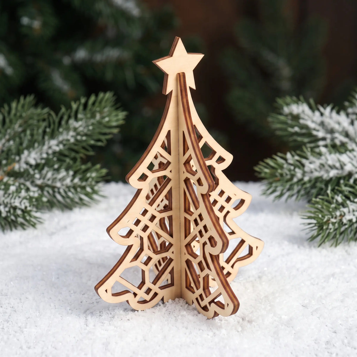 3D laser cut plywood Christmas tree figurine standing on white artificial snow with frosted pine branches in background.