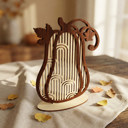 A tall, slender decorative pumpkin made of laser cut plywood featuring a modern arch and stripe pattern. The dark wood silhouette contrasts with the light plywood interior. Shown standing on a table with scattered yellow maple leaves.