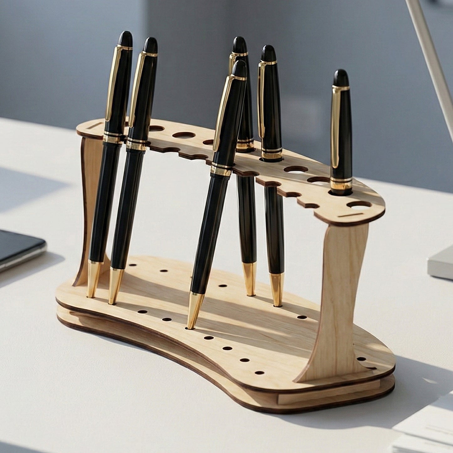 Minimalist laser cut plywood pen stand with a double-deck arc design. Features ten circular slots to hold pens vertically. Shown on a white office desk with black and gold pens, a smartphone, and documents to demonstrate scale.