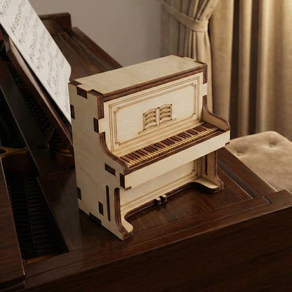 Miniature upright piano secret box made of laser cut plywood, resting on a full-sized dark wood piano. The box features an engraved music book and detailed piano keys. Warm interior lighting and sheet music in the background provide a musical context