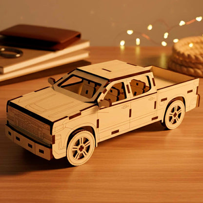 Laser cut wooden plywood 3D puzzle of a pickup truck. Detailed front view showing the grill, headlights, and bumper. The model is made of light plywood with dark burnt edges, resting on a wooden table next to books and leather notebooks.