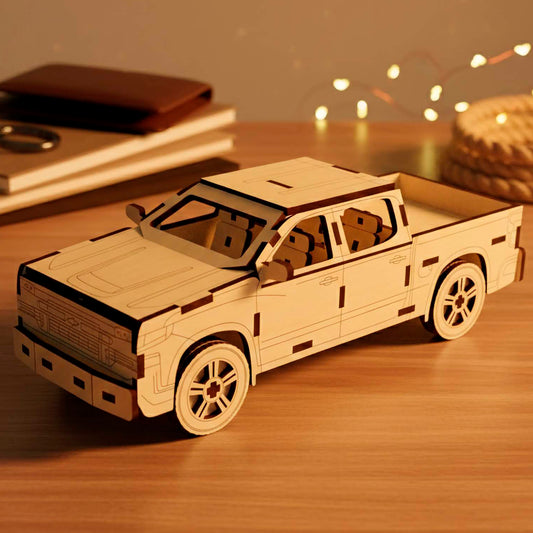 Laser cut wooden plywood 3D puzzle of a pickup truck. Detailed front view showing the grill, headlights, and bumper. The model is made of light plywood with dark burnt edges, resting on a wooden table next to books and leather notebooks.