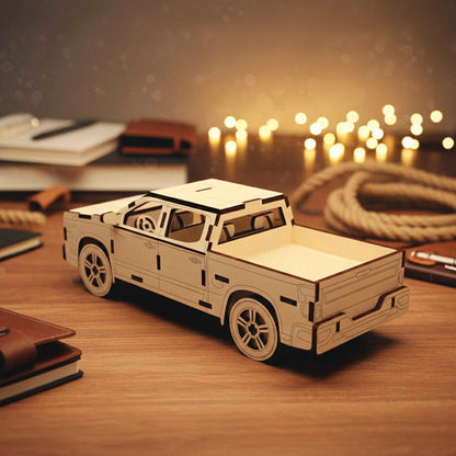 Rear perspective of a laser cut wooden plywood pickup truck model. Shows the open truck bed, detailed tailgate, and layered wheels. The light wood craft is displayed on a desk with a rope and bokeh lights in the background.
