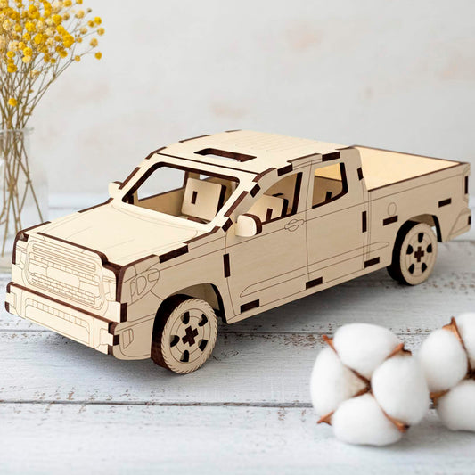 A laser cut wooden  plywood pickup truck model shown from an elevated front angle. The 3D wooden toy features intricate interior seating and engraved details, set in a bright room with yellow flowers and cotton bolls on a white wooden surface.